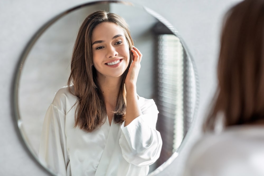 Woman smiling at herself in the mirror, happy with the results from her Juvederm treatment.